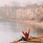 Der Blick erstreckt sich über von der Klippe über den See bis zu endenen Steinwand, während die Sonne aufgeht streckt sich eine Frau ihr entegegen.